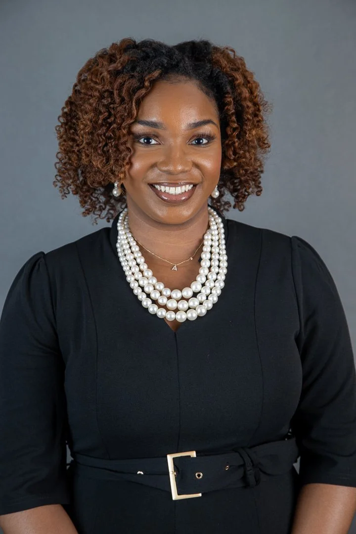 A woman with curly, shoulder-length hair wearing a black dress with a belt, layered pearl necklaces, matching pearl earrings, and a gold necklace with a small triangle pendant, smiling against a gray background.