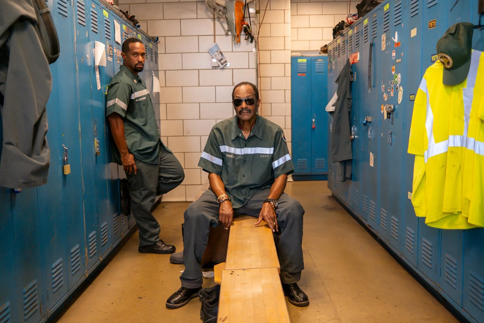 Three men in gray uniforms inside a locker room. One man is sitting on a wooden bench, wearing sunglasses. Two others are standing, one leaning against lockers, in a serious pose. The locker room has blue lockers with various stickers and personal it
