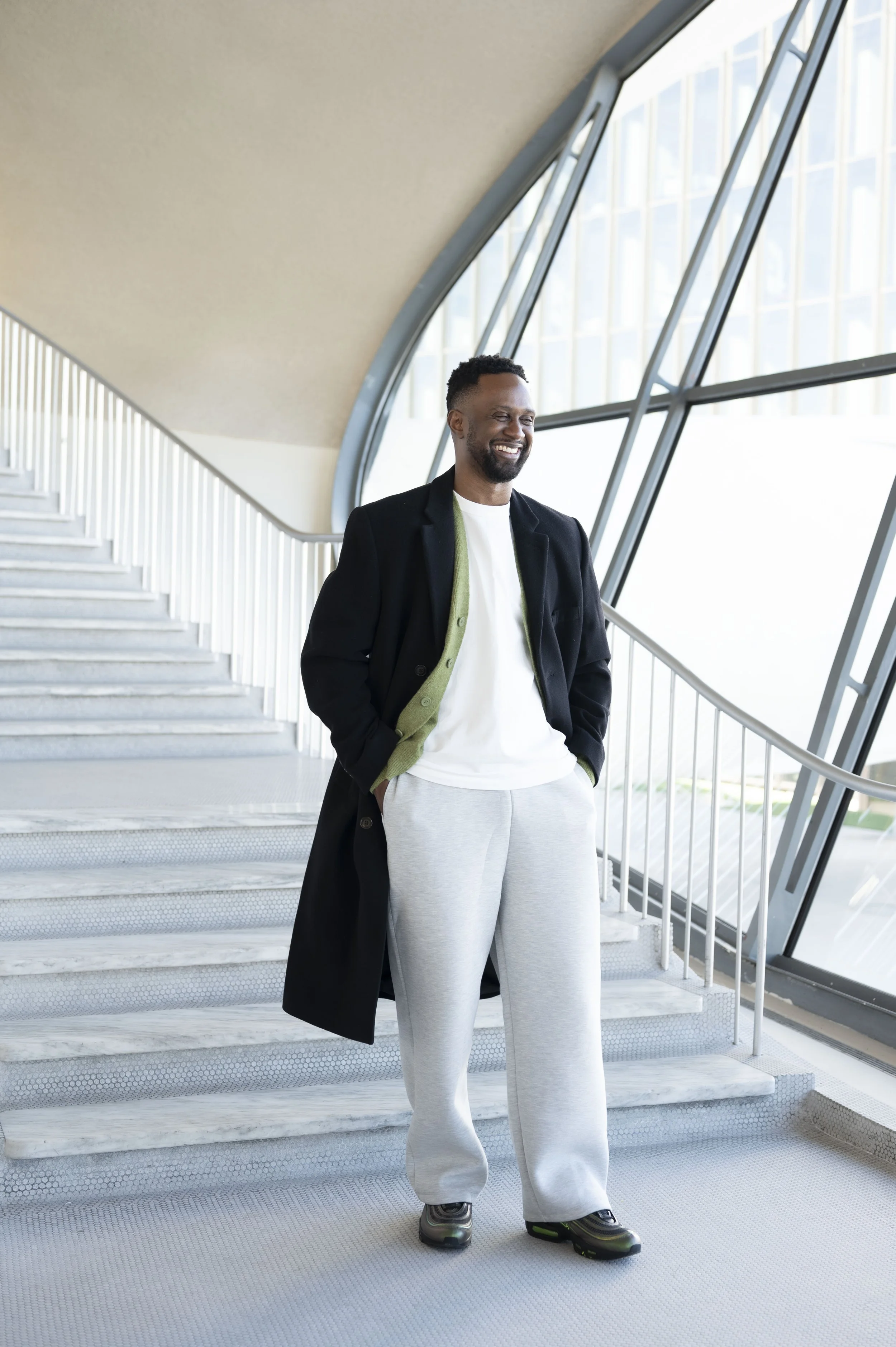 A smiling man in casual clothes standing inside a modern building, near a staircase and large glass windows with steel framing.