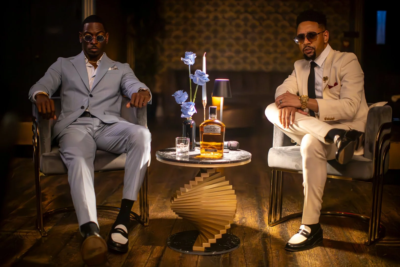 Two stylish men sitting on armchairs in a dimly lit room with a decorative table between them, featuring a bottle of alcohol, blue flowers, a lit candle, and glassware.