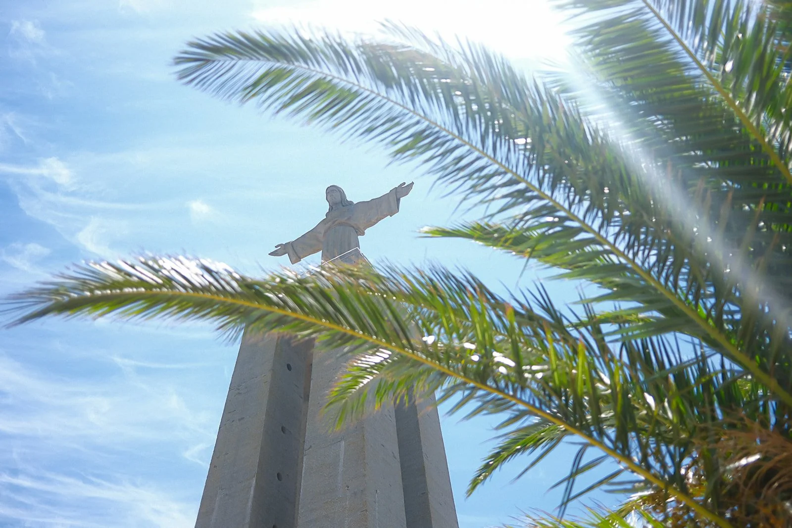 Close-up of a statue of Jesus Christ with outstretched arms seen through palm tree leaves against a blue sky.
