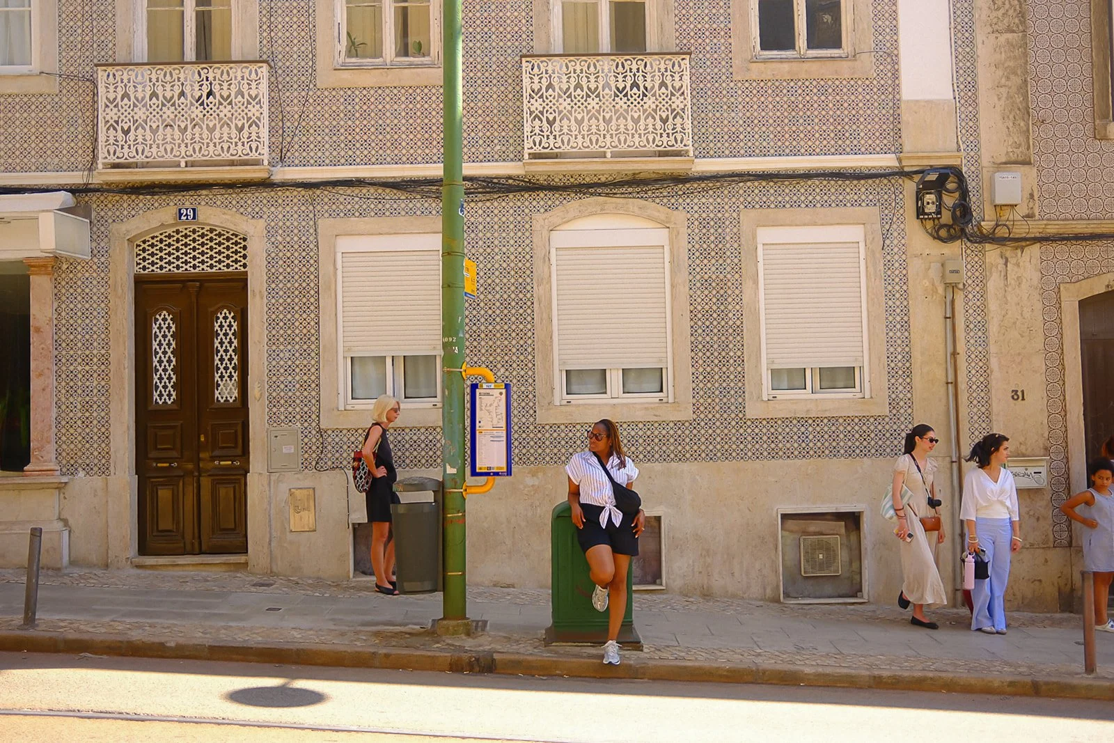 People standing and sitting on a sidewalk in front of a building with windows and decorative tilework. There is a woman with white hair, sunglasses, and a black dress standing near a door, another woman with sunglasses leaning against a green post, a
