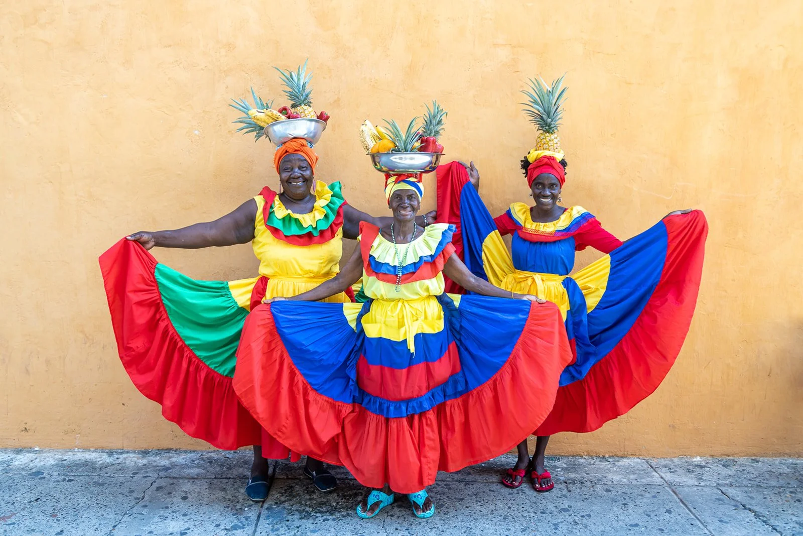 Three women dressed in colorful traditional costumes, holding out their skirts, and balancing bowls of fruit on their heads, standing against a yellow wall.
