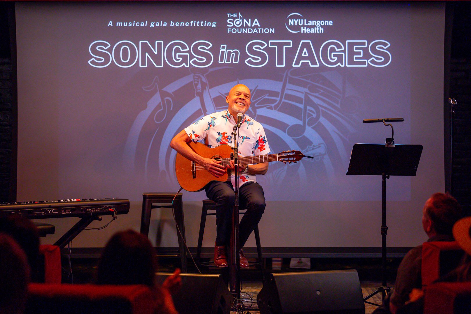 A man wearing a floral shirt is sitting on a stool, playing an acoustic guitar and singing into a microphone on a stage. Behind him, a large screen displays text for a benefit event called 'Songs in Stages,' with musical notes in the background and l