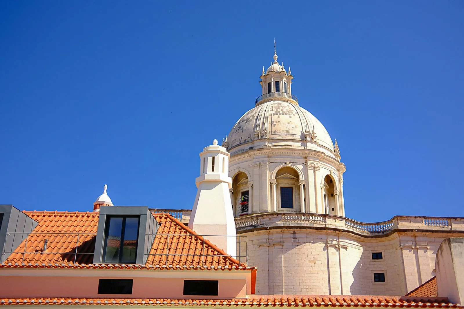 View of a historic building with a large, rounded dome and smaller ornate details, with a modern building with large windows and a red-tiled roof in front, under a clear blue sky.