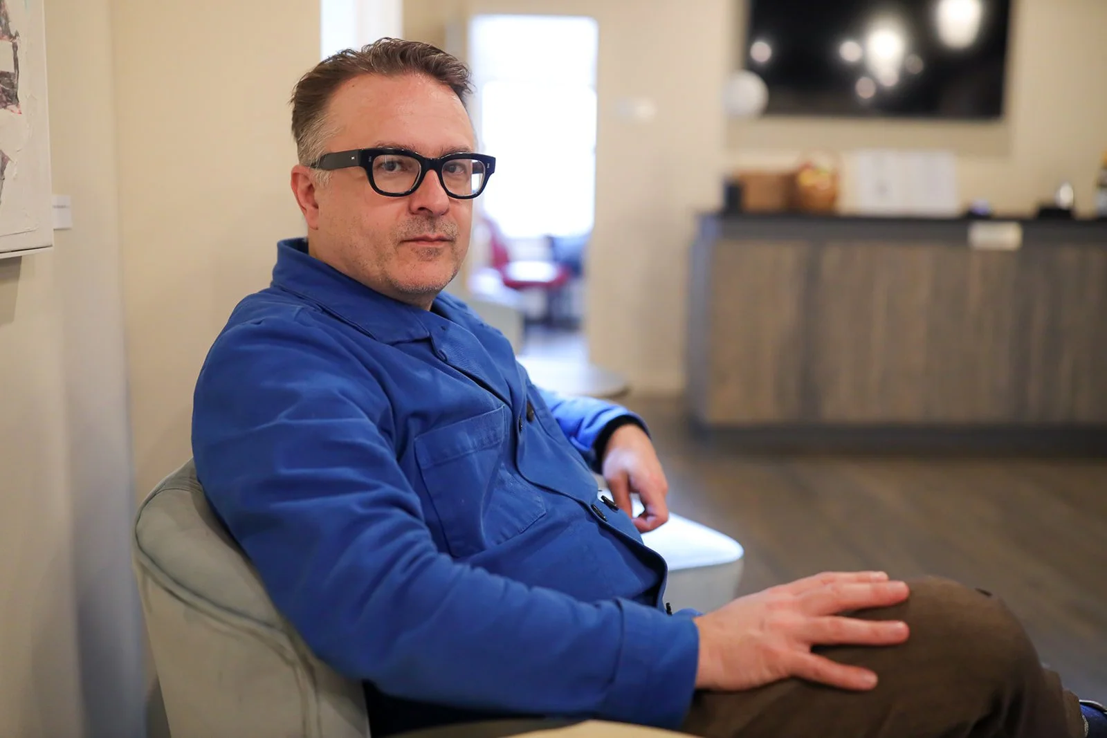 A man with glasses and a blue jacket sitting on a beige chair in a room, looking at the camera with a neutral expression.