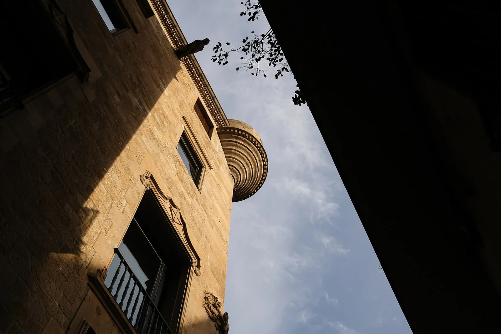 Looking up at a historic stone building with ornate window frames and a rounded corner turret, partially shaded, with a small tree branch and sky in the background.