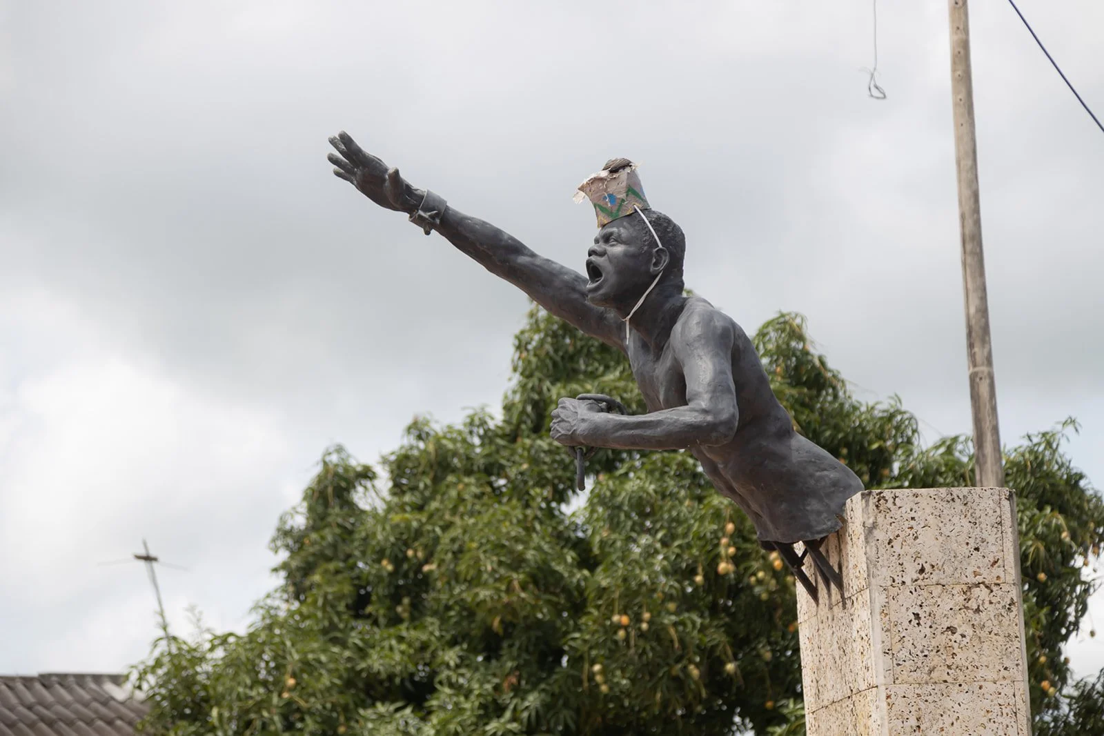 A bronze statue of a man with a crown, shirtless, with his right arm extended forward and left arm bent holding an object, mounted on a stone pedestal against a cloudy sky and trees.