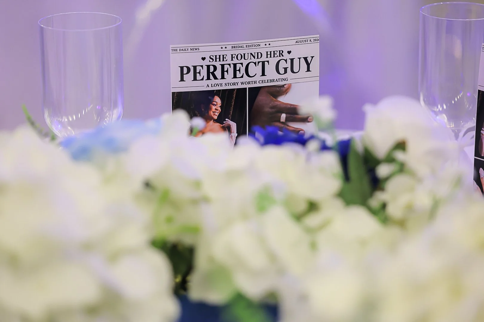 A wedding reception table with a newspaper headline, two empty wine glasses, and a floral arrangement in the foreground.