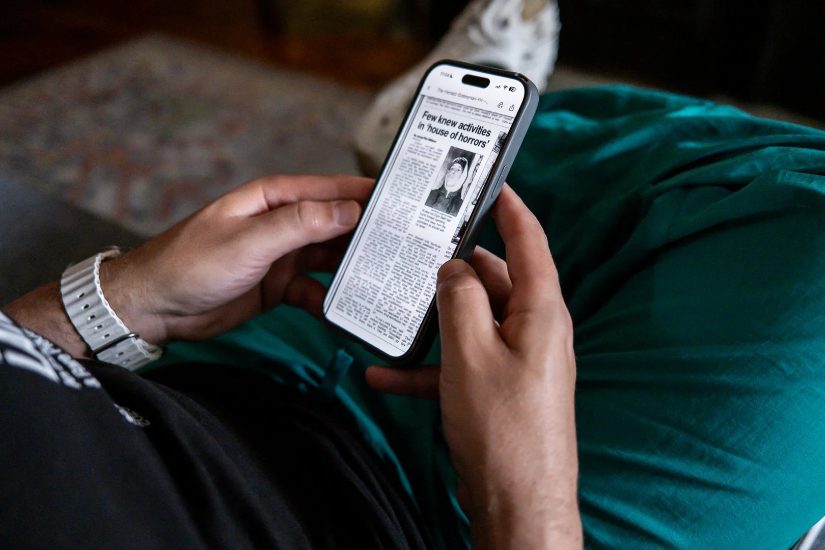 A person holding a smartphone reading a newspaper article about a house of horrors, with a black and white photograph of a person in the article.