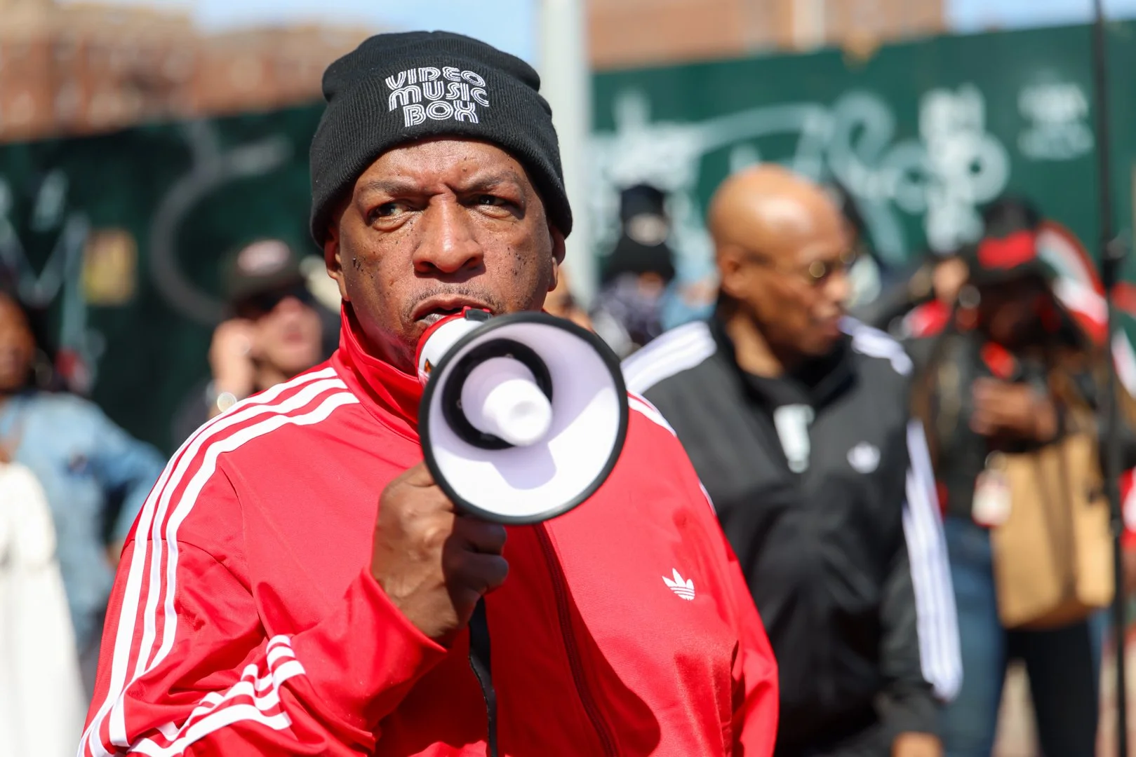 A man wearing a black beanie with the words 'Video Music Box' holding a megaphone, surrounded by a crowd in an outdoor setting.