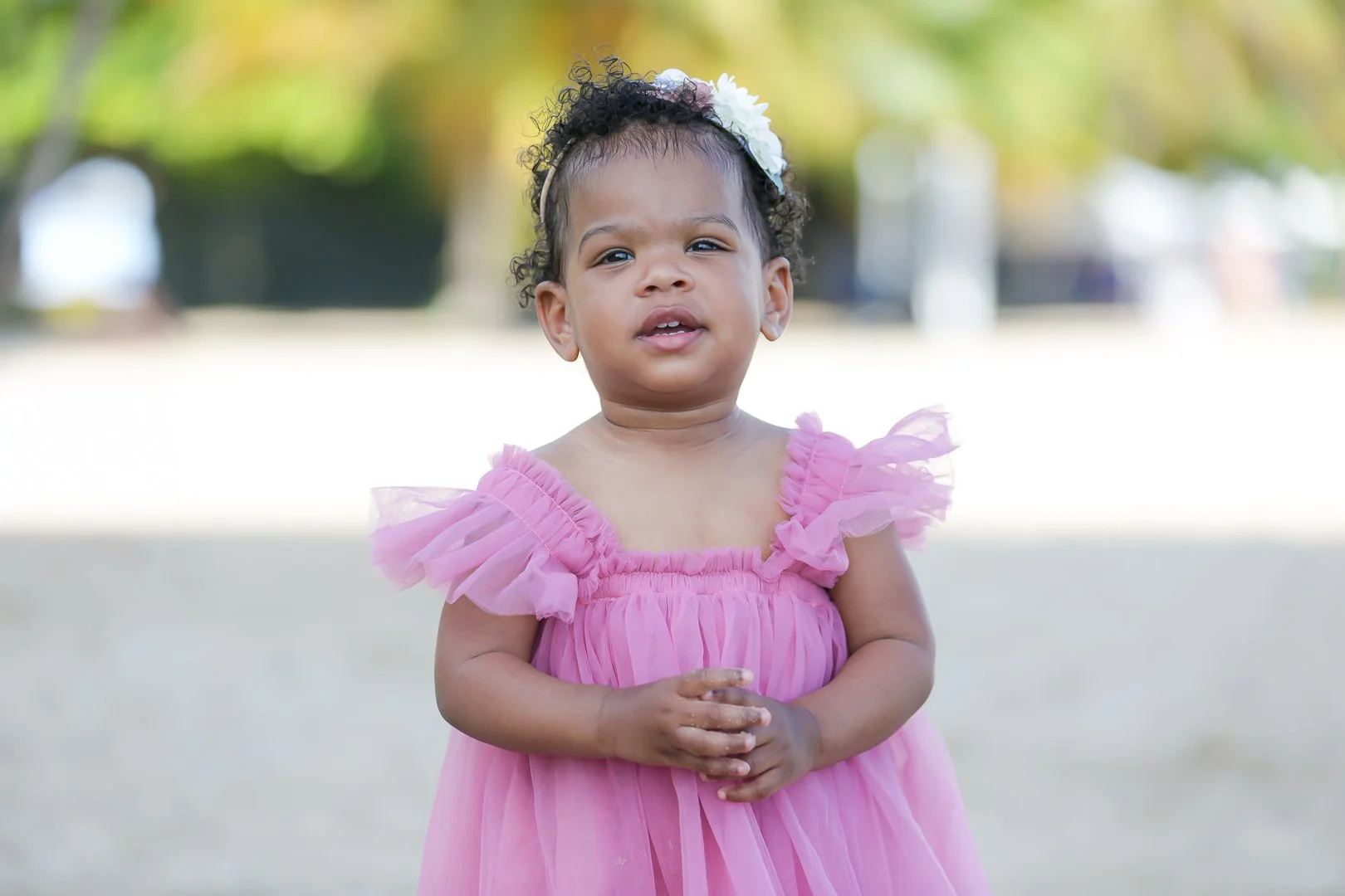 A young girl with curly hair wearing a pink dress with ruffled sleeves and a white floral headband, standing outdoors with blurred trees in the background.