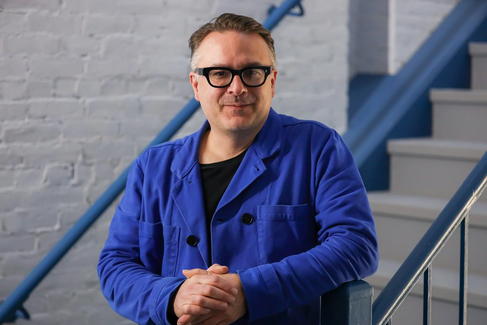 A man with glasses and short brown hair, wearing a blue jacket, standing on a staircase with a white brick wall in the background.