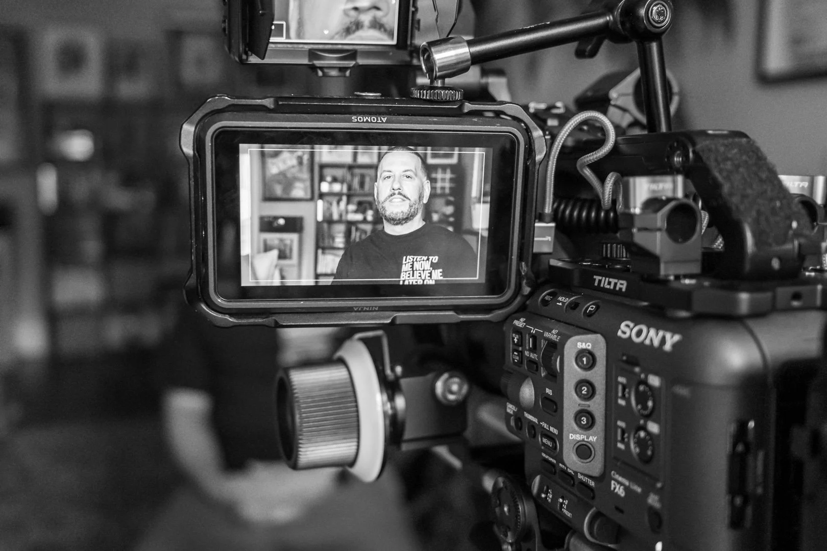 Close-up of a professional Sony video camera capturing a man speaking during an interview, with a bookshelf in the background.