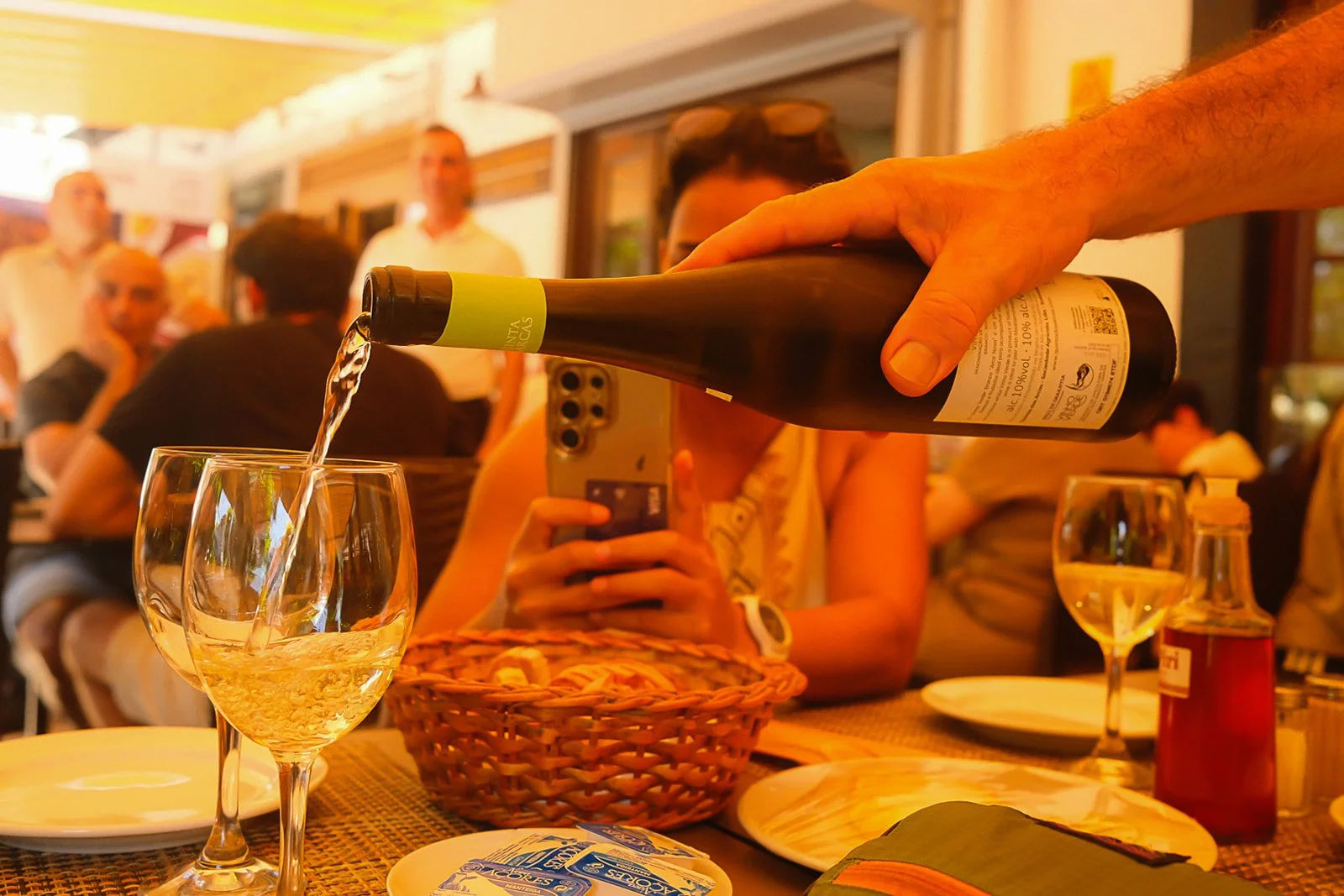 A person pouring white wine into a glass at a restaurant table with several other glasses, plates, and people in the background.