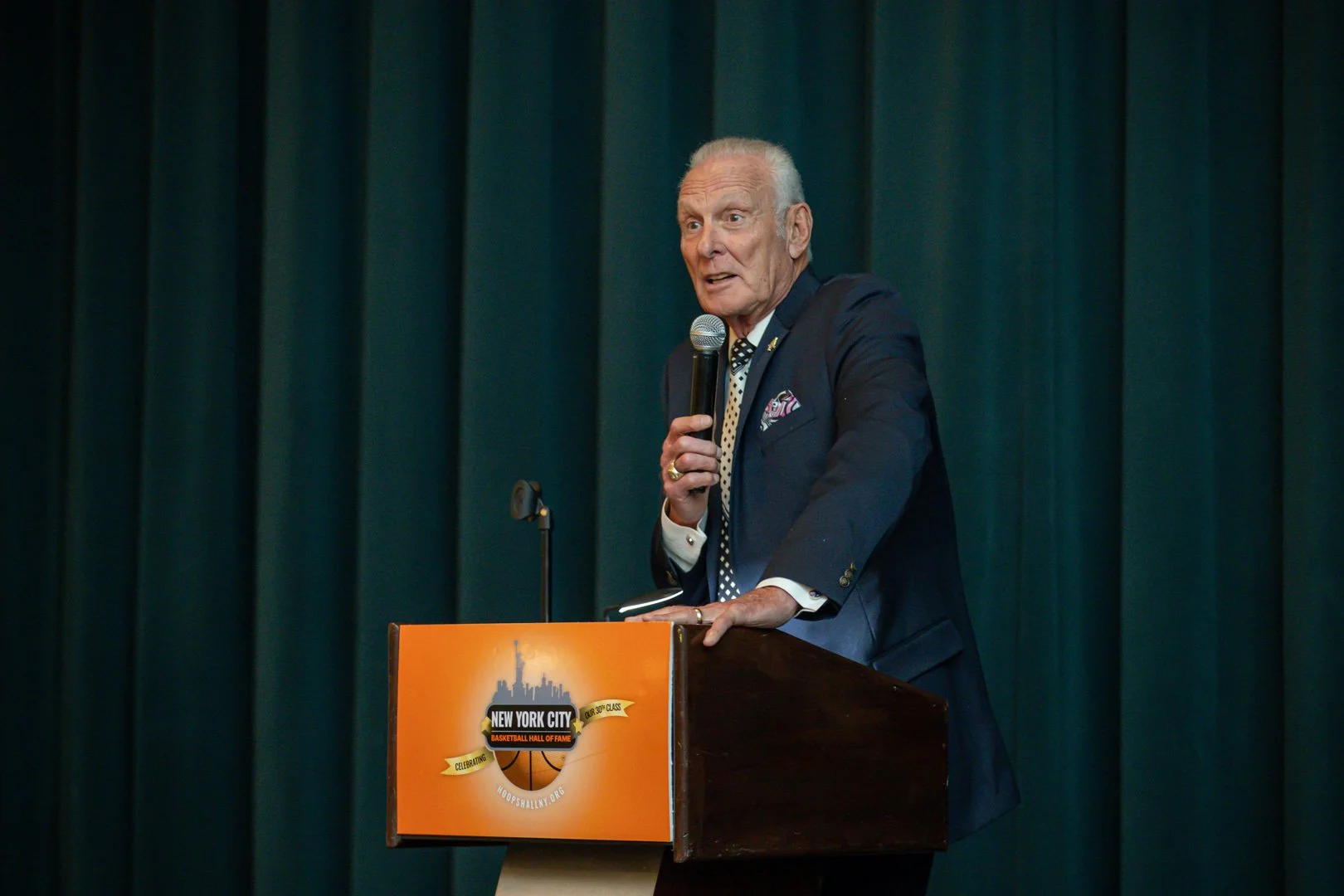 An elderly man speaking into a microphone at a podium during an event honoring the Basketball Hall of Fame in New York City, with dark green curtains in the background.
