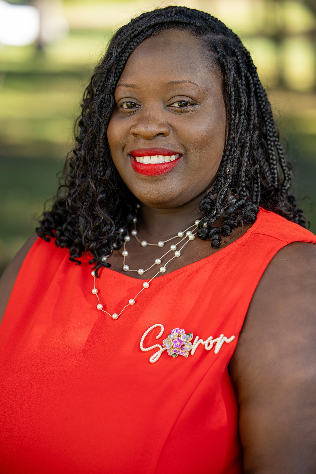 A woman with dark braided hair and bright red lipstick smiling, wearing a red sleeveless top with a flower and the word 'Sorry' embroidered on it, and layered pearl necklaces, standing outdoors with a blurred green background.