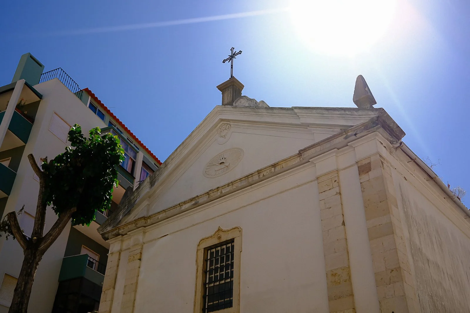 A small white church with a cross on top, against a bright blue sky, with a modern apartment building nearby and a tree in the foreground.