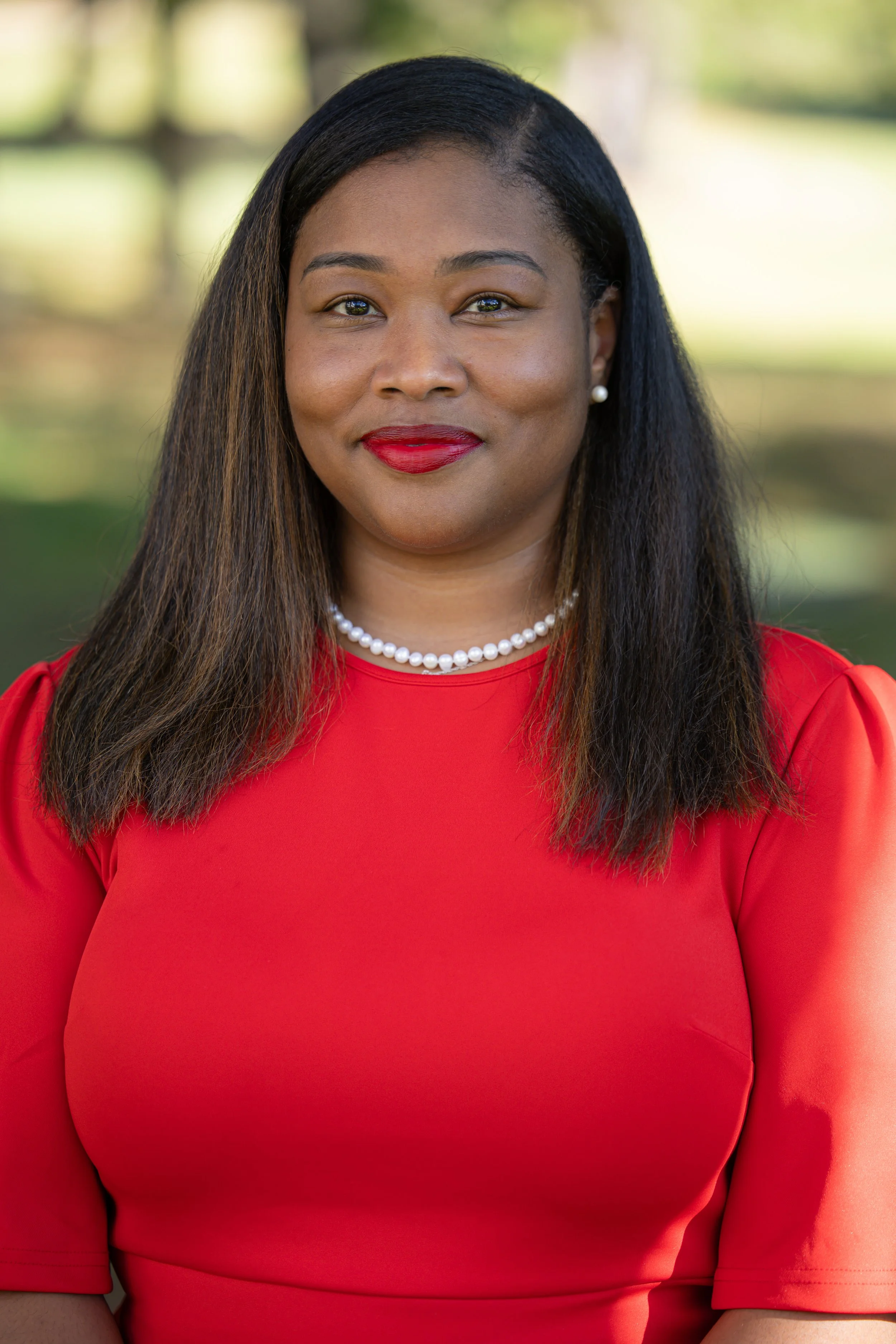 A woman with medium brown skin, long dark hair, wearing a red dress, pearl necklace, and pearl earrings, standing outdoors with blurred greenery in the background.