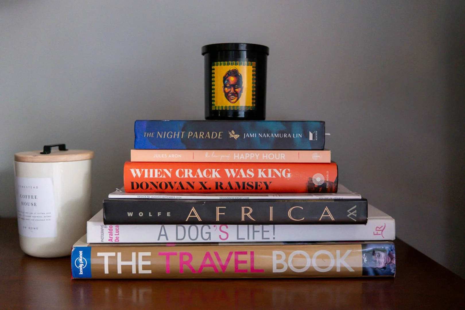 A stack of six books on a table with a container to the left. The titles include "The Night Parade," "Happy Hour," "When Crack Was King," "Africa," "A Dog's Life!" and "The Travel Book." A black container with an artistic image of a woman’s face sits