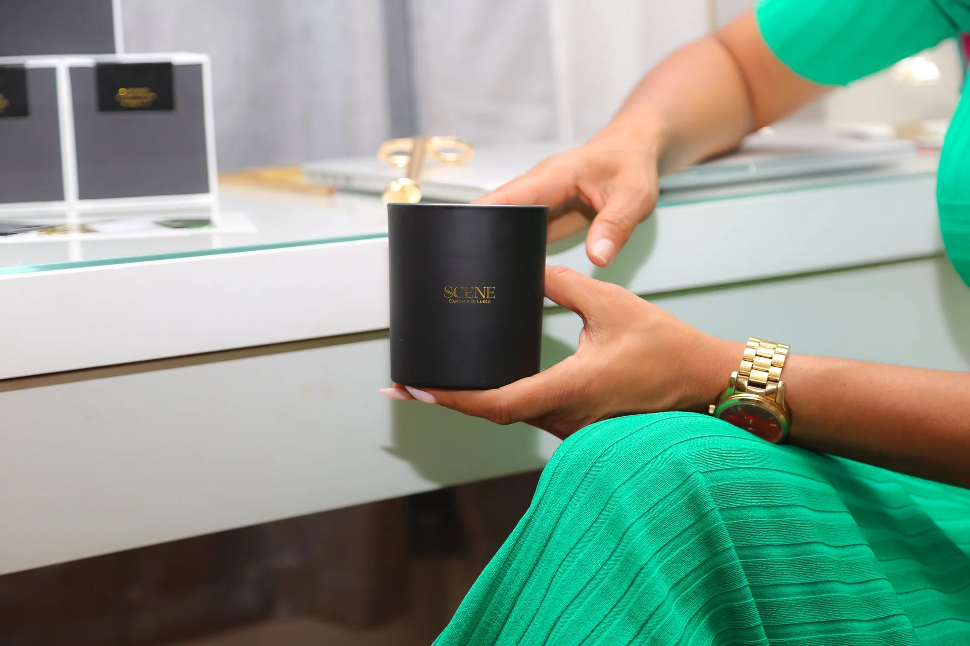 A woman wearing a gold watch and green pants is holding a black candle with gold lettering that says 'SCENE CANDELE DI LUSSO' while sitting in front of a display counter with other black candles.