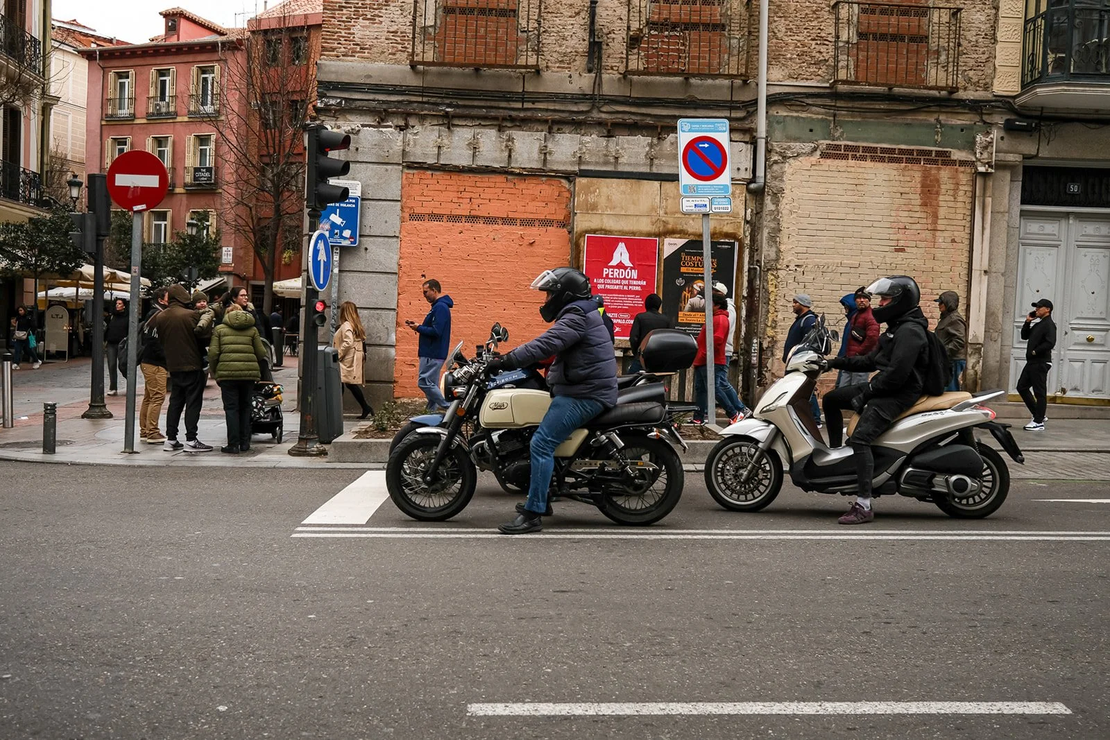 Two motorcyclists waiting at a city crosswalk, with a group of pedestrians on the sidewalk behind them. The scene captures an urban street with various signs and a partially building wall.