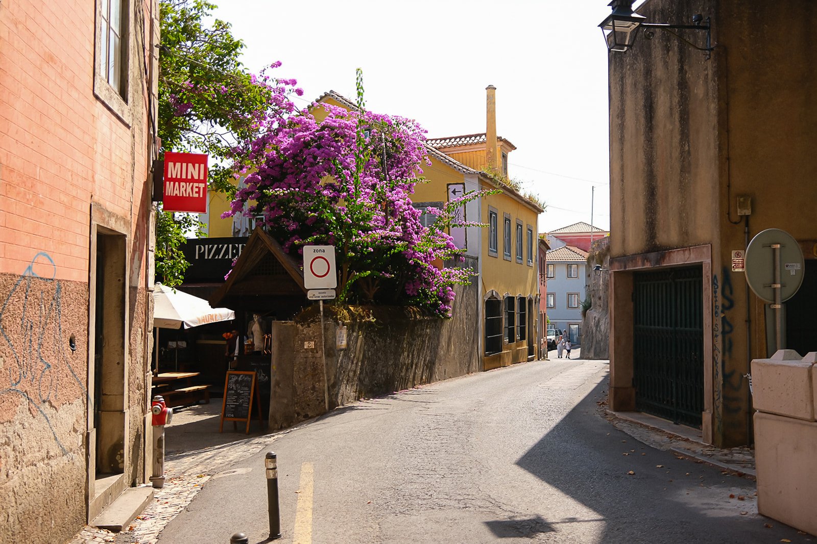 A narrow street in a European town with colorful buildings, a large pink flowering tree, signs for a mini market, pizzeria, and a no-entry zone, with pedestrians in the distance.