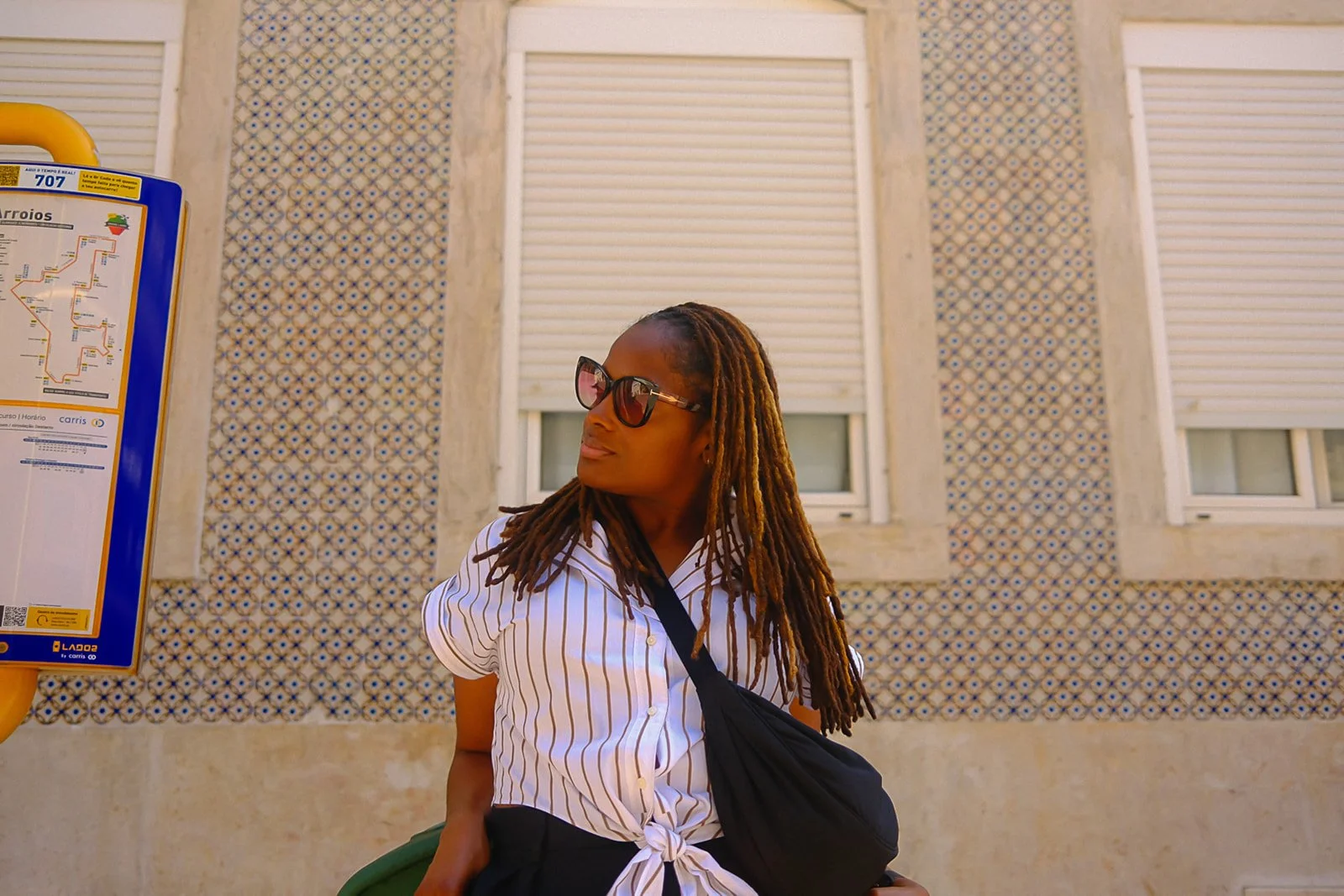 A woman with long dreadlocks wearing sunglasses, a white and beige striped shirt, and a black bag, standing at a bus stop in front of a textured wall with three windows, and a bus schedule sign on the left.