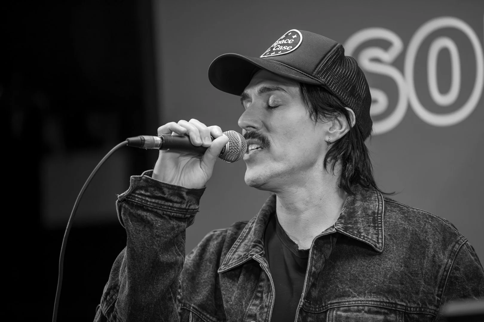 A man singing into a microphone with eyes closed, wearing a trucker hat and denim jacket, in front of a background with the word 'SO'.