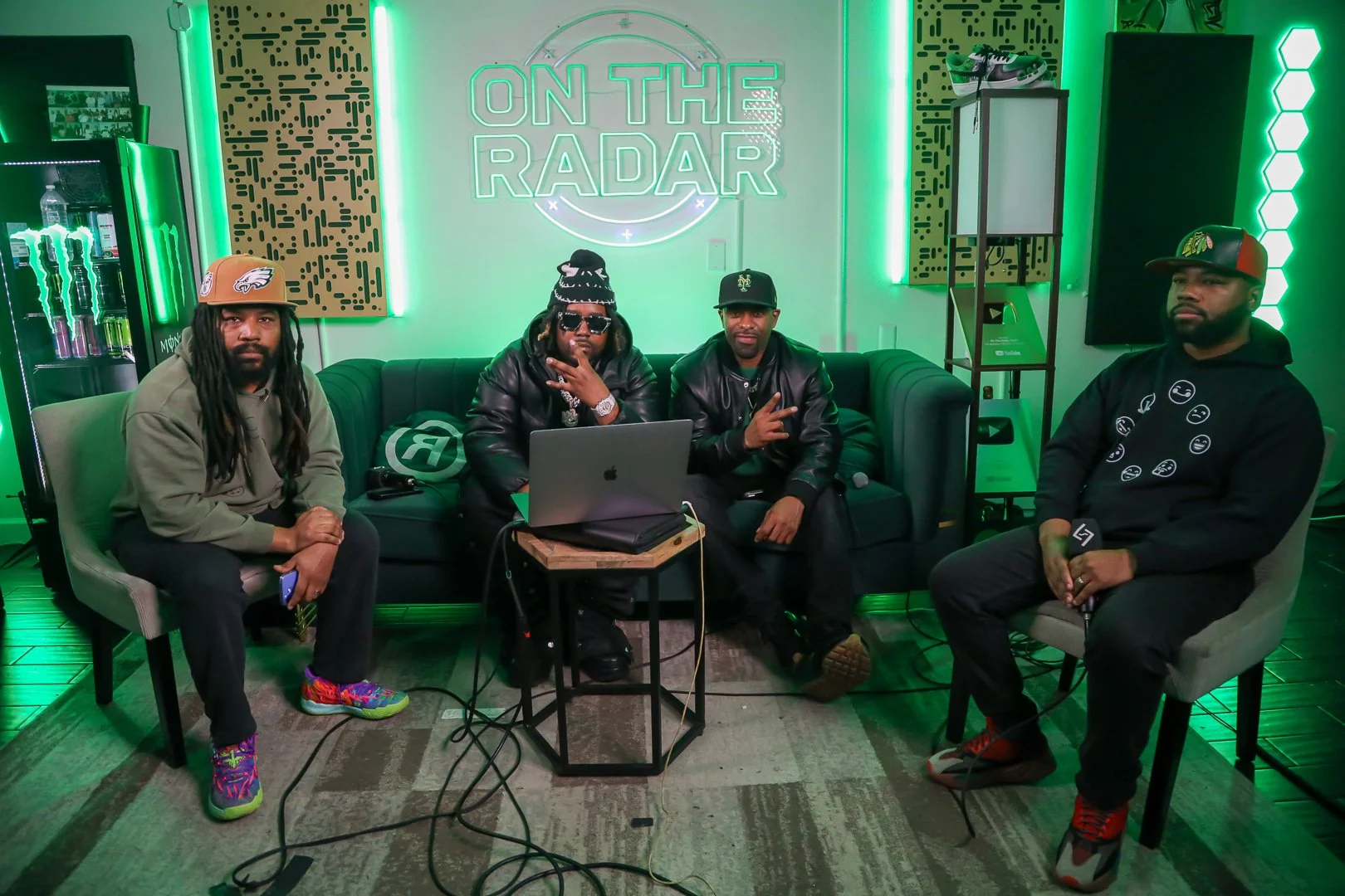 Four men sitting in a room with green neon lights, two seated on a black sofa and one on grey armchairs. They are in a room labeled 'On The Radar,' with audio and recording equipment around them.