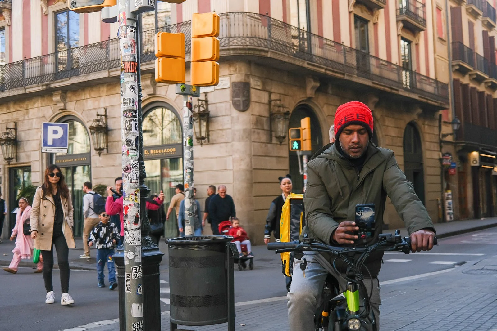 Man wearing red beanie riding a bicycle on city street while pedestrians walk in the background near a corner building with ornate architecture and street signs.