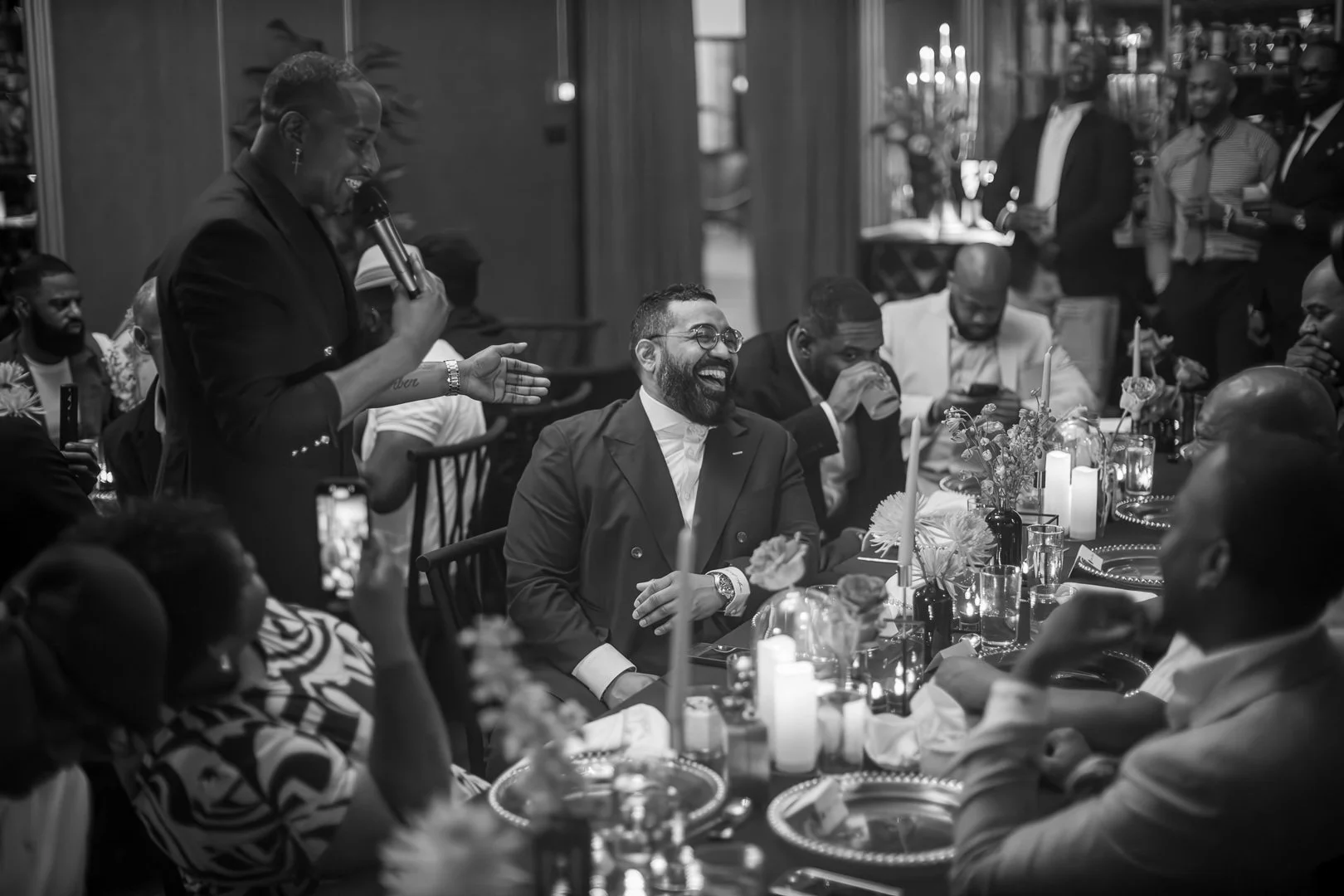 A group of people at a formal dinner event, with some individuals laughing and engaging in conversation. The scene is in black and white, with a woman standing and speaking into a microphone.