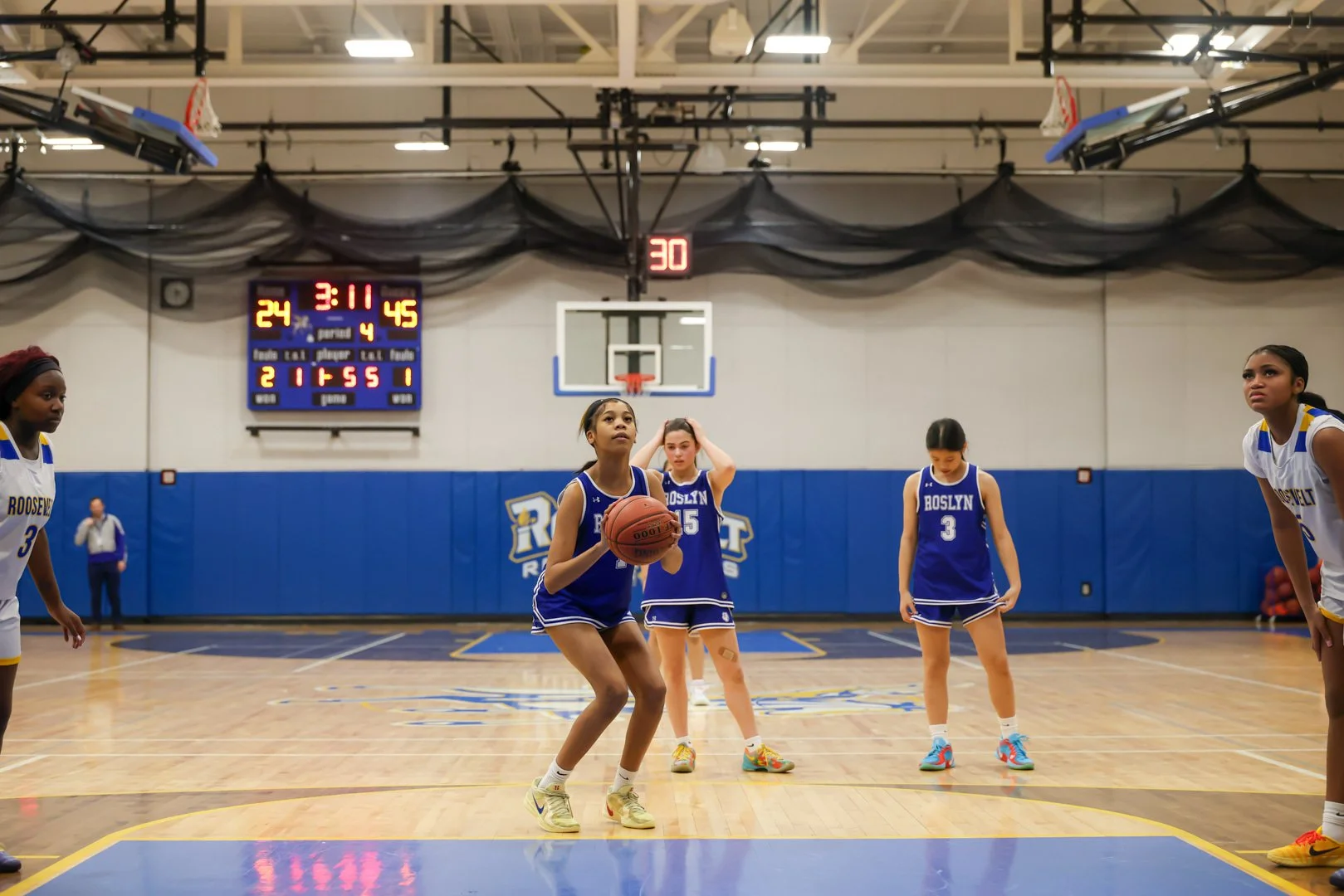 Girls' basketball game in an indoor gym, with one girl preparing to shoot free throws and others standing nearby, score and time displayed on scoreboard.
