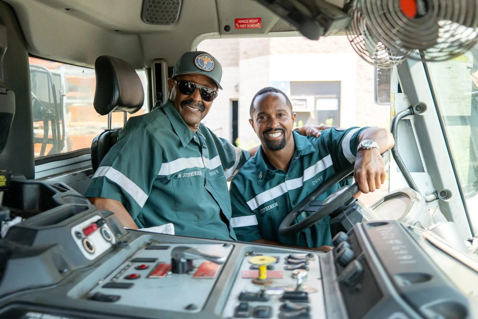 Two smiling men wearing green work uniforms inside a vehicle, with one sitting in the driver's seat and the other standing beside him, showing camaraderie.