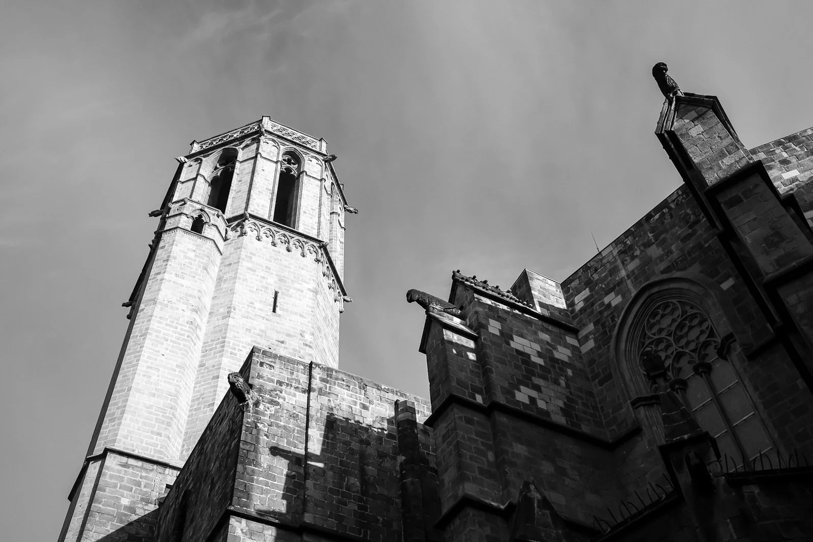 Black and white photo of a tall church tower and part of the church building with a Gothic-style arched stained glass window, with the sky in the background.