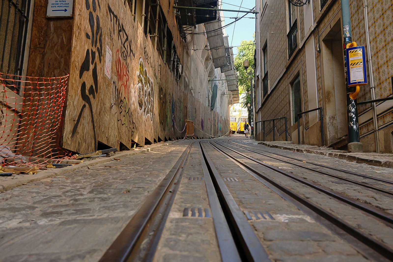 Street view with tram tracks running through a narrow, cobblestone alleyway. On the left, there is a construction area enclosed by a plywood barrier and orange netting. On the right, there are buildings with decorative tiles and a public phone booth 