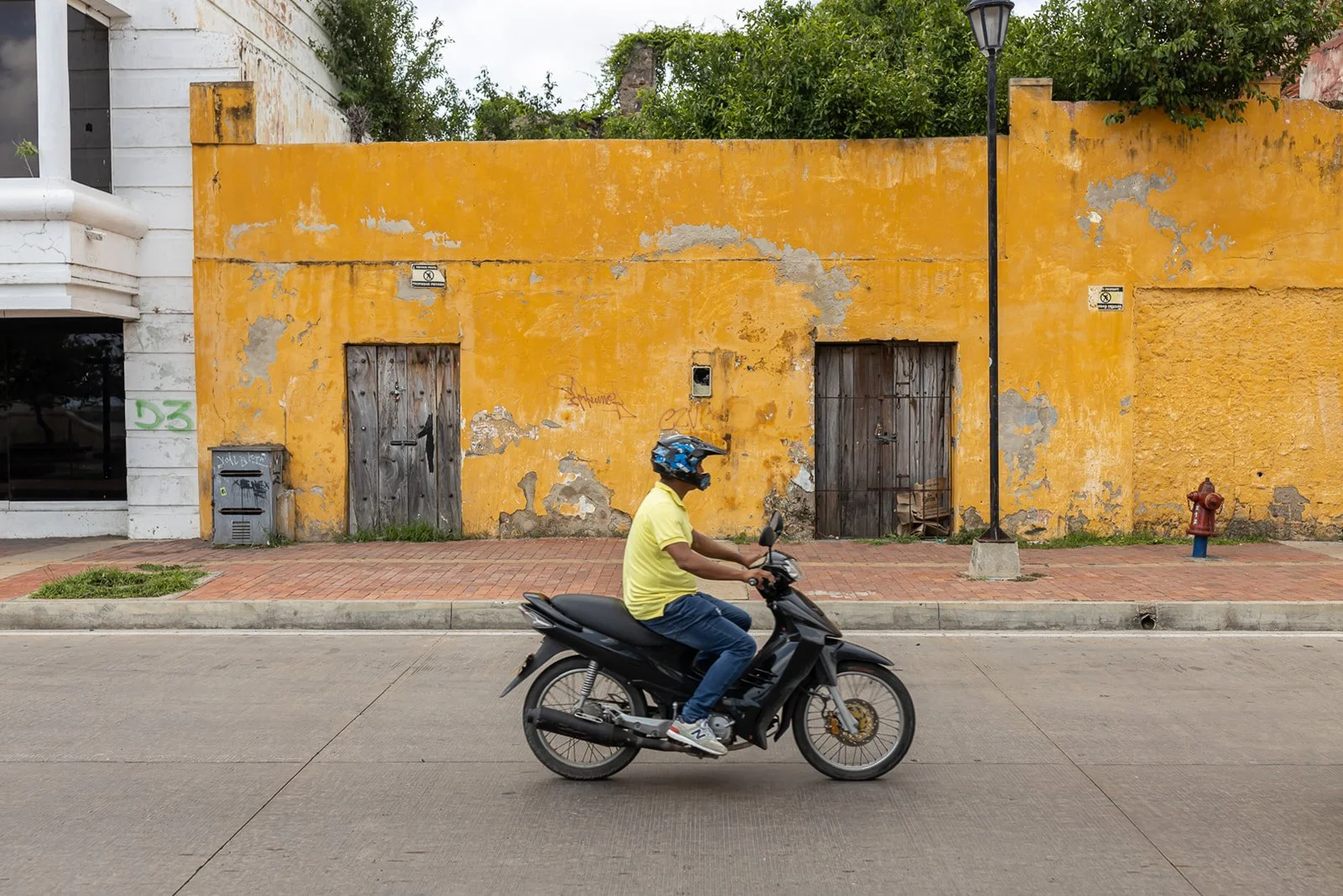 A man wearing a yellow shirt, jeans, and a helmet rides a motorcycle on a city street in front of a weathered yellow wall with two wooden doors, graffiti, and a small mailbox. A street lamp and fire hydrant are also visible.