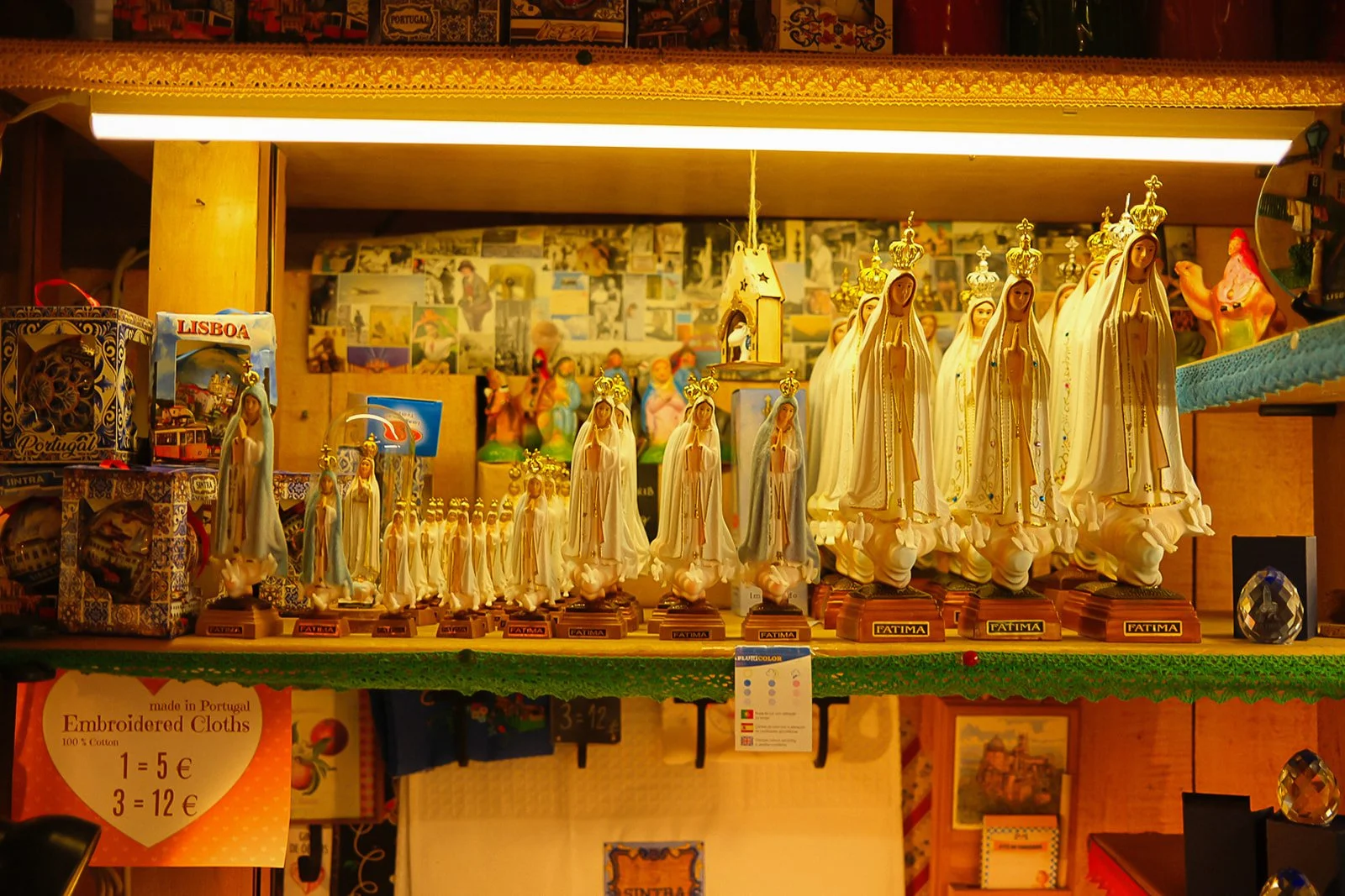 Shelf displaying various religious statues of the Virgin Mary and other figures, with small souvenir boxes, framed pictures, and posters in the background.