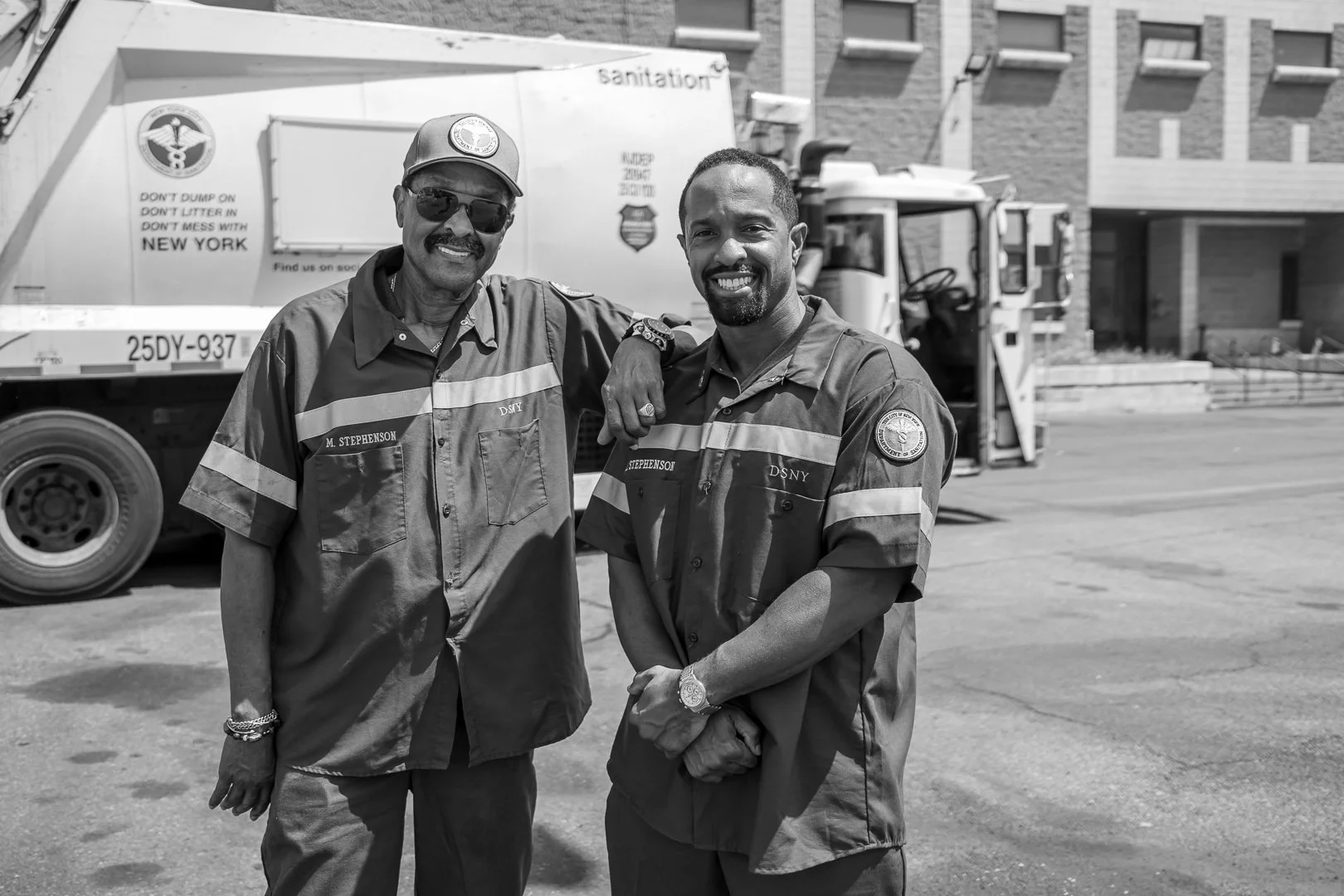 Two men dressed in New York City sanitation worker uniforms standing in front of a sanitation truck, smiling at the camera.