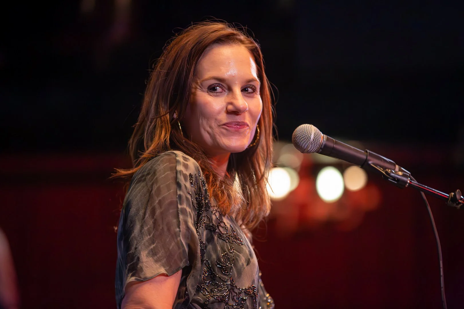 A woman with shoulder-length brown hair and hoop earrings is smiling at the camera while standing next to a microphone on a dark stage with blurred lights in the background.