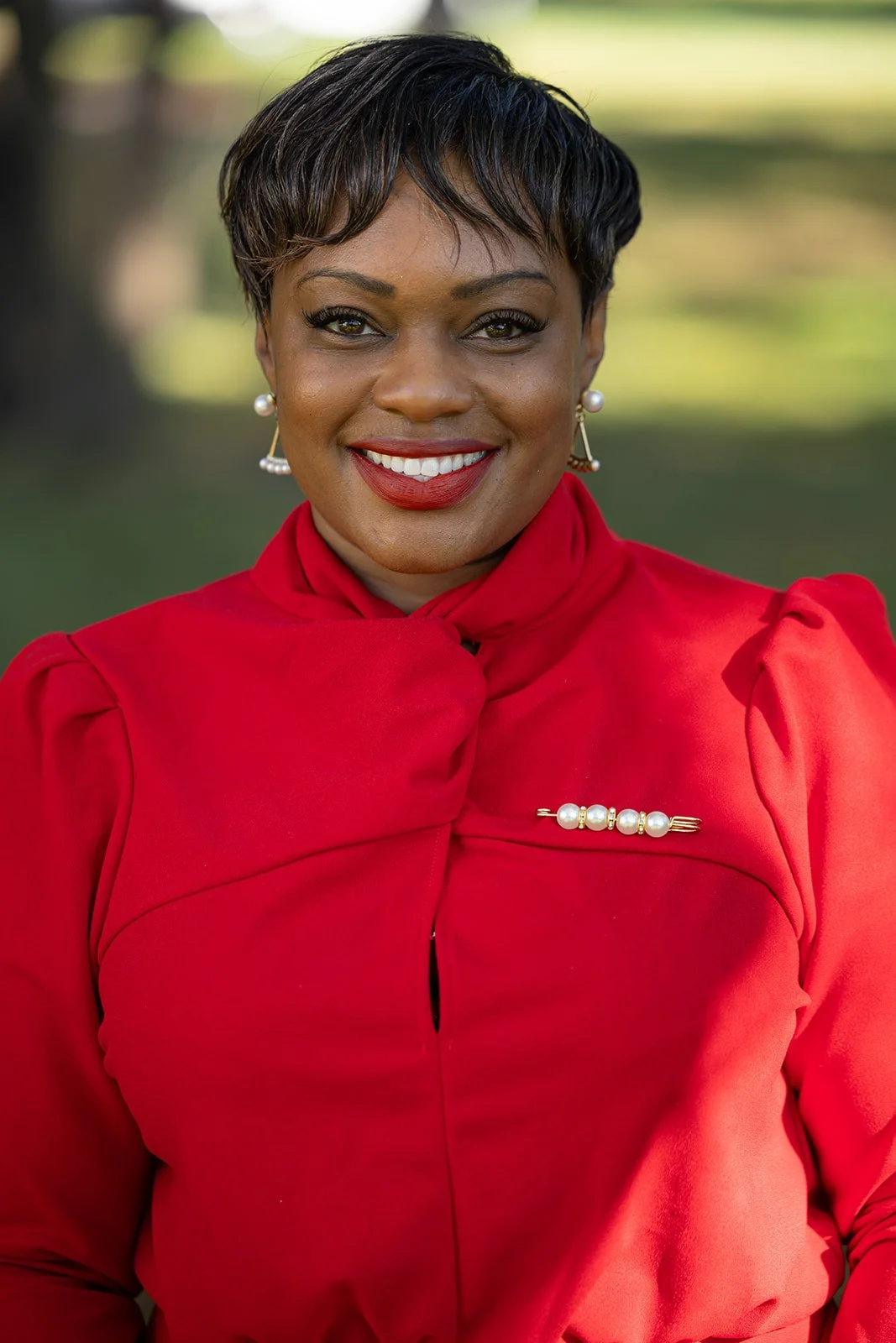 A woman with short black hair, wearing a red dress with puffed shoulders and pearl earrings, smiling outdoors with a blurred green background.