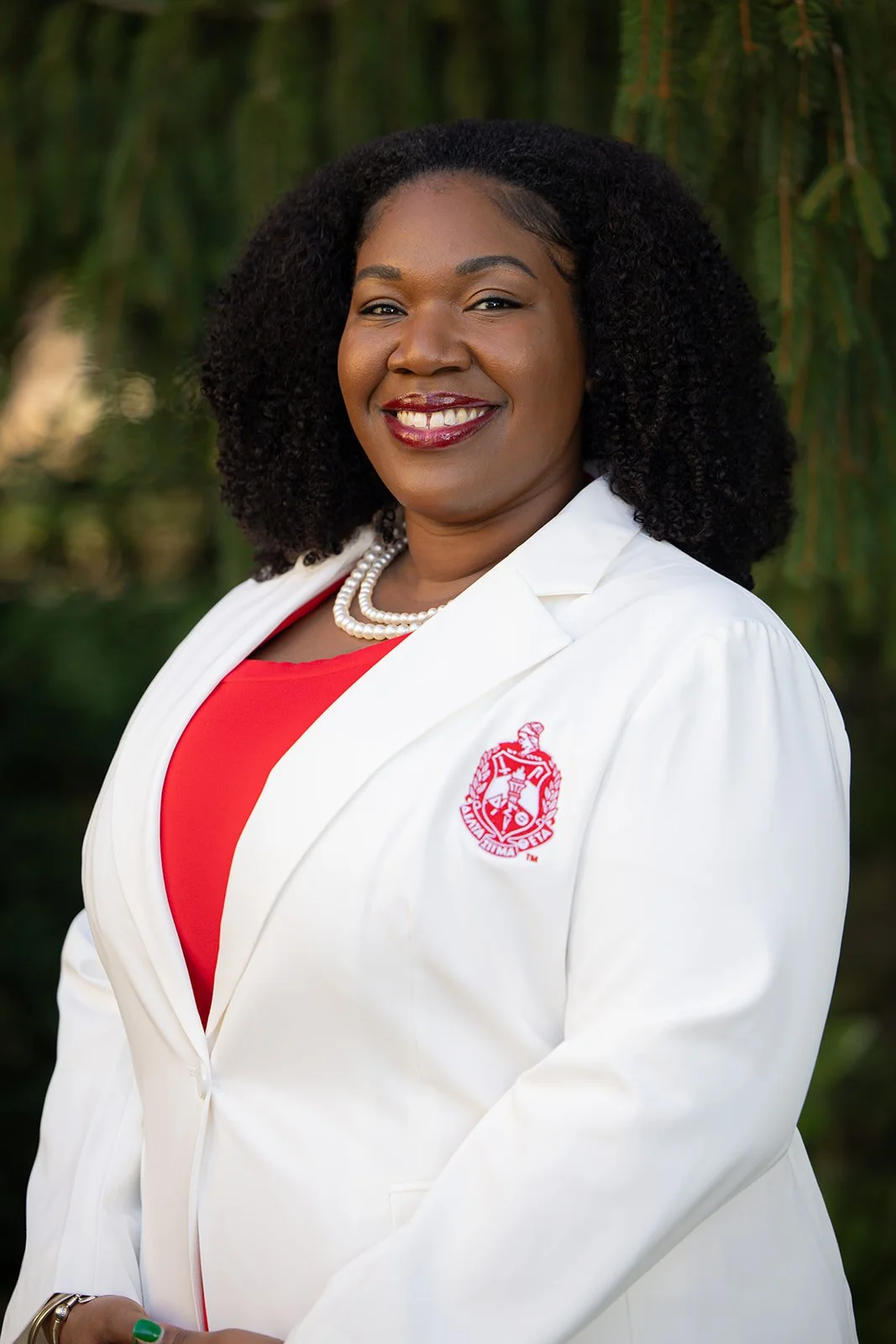 A woman with dark curly hair smiling, wearing a white blazer with a university logo, a red top, a pearl necklace, and standing outdoors with greenery in the background.