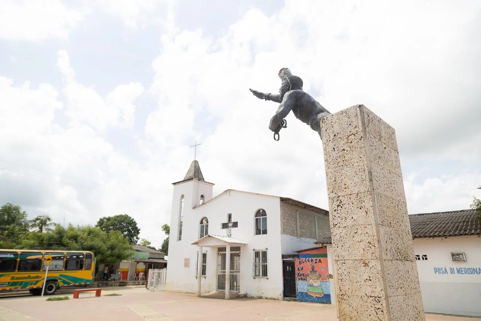 Statue of a soldier leaning forward, mounted on a tall stone pedestal, in front of a white church with a cross on its steeple; cloudy sky and trees in background, yellow bus and posters on wall.