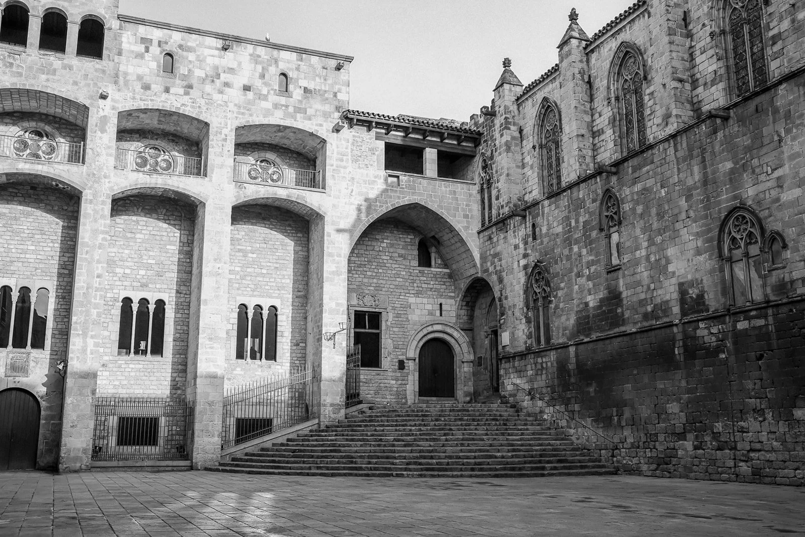 Black and white photo of an old stone building with arched windows and doors, stone stairs leading up to a doorway, and Gothic-style windows on the right side.