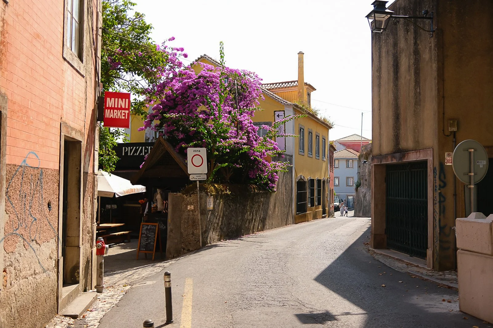 A narrow street in a European town with colorful buildings, a blooming purple bougainvillea, signs for a mini market and pizzeria, and a few pedestrians in the distance.
