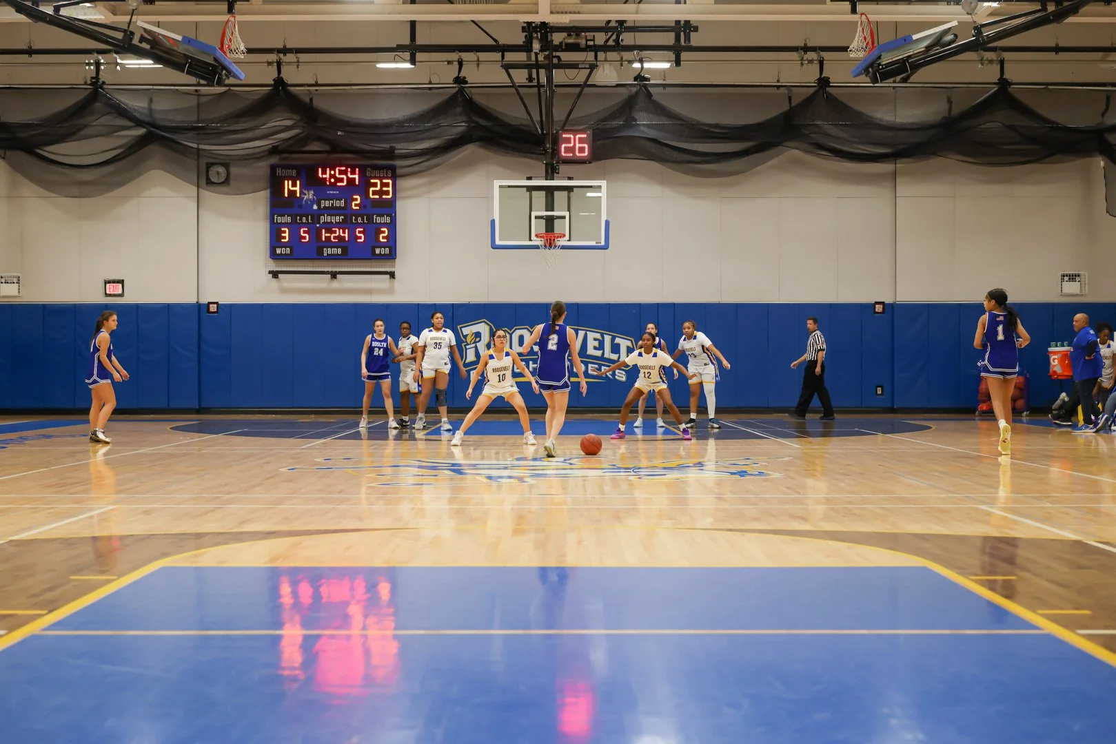 Young female basketball players on a gym court preparing for a free throw shot during a game, with a scoreboard showing 14-23 and 4 minutes and 54 seconds remaining.