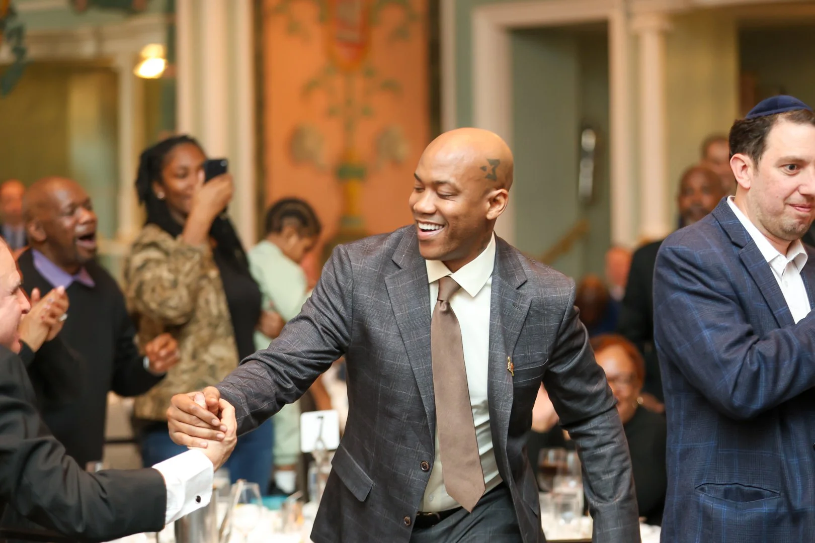 A man in a brown plaid suit shaking hands with someone at a formal event or celebration, smiling happily. Other attendees are seen clapping, laughing, and taking photos in the background.