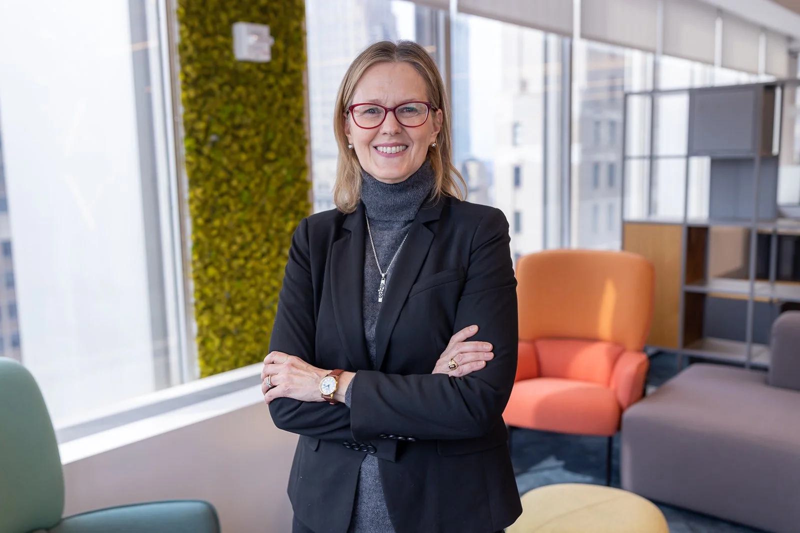 A professional woman with shoulder-length blond hair, red glasses, and pearl earrings, standing with arms crossed in a modern office with large windows, colorful chairs, and bookshelves.