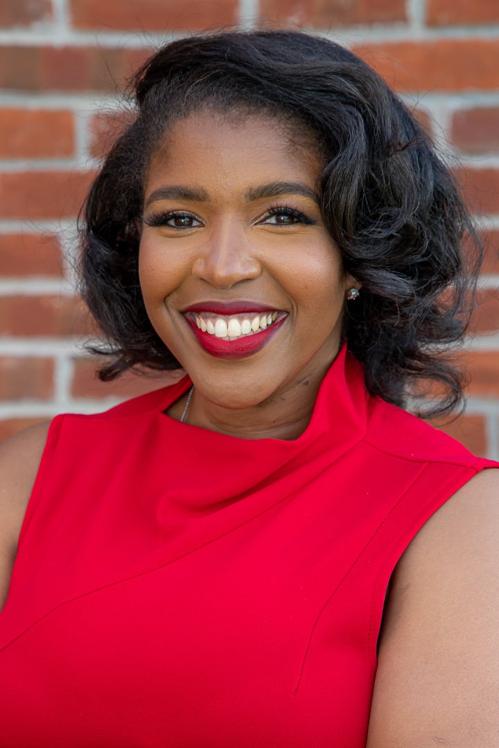 A woman with dark hair styled in loose waves, wearing a red sleeveless top, smiling, standing in front of a brick wall.