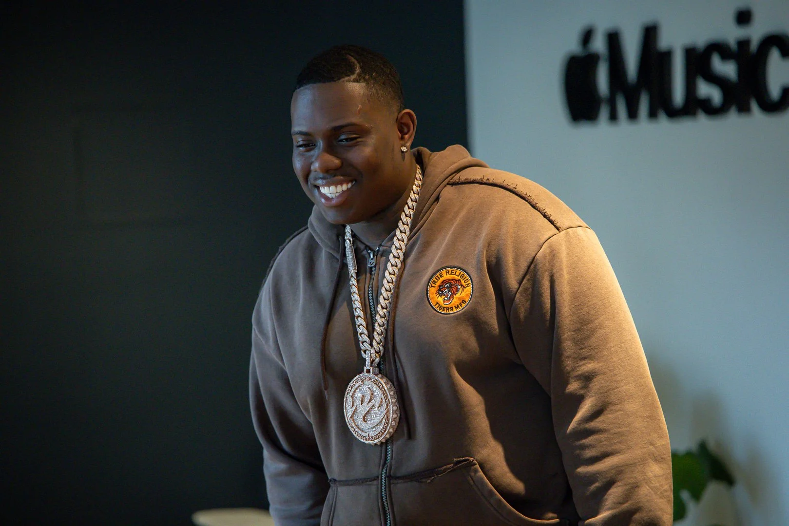 A smiling young man wearing a brown hoodie with a Tiger logo patch, gold chain with a large pendant, and earrings, standing in an indoor setting with a sign that reads 'iMusic' in the background.